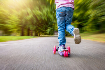 Back view of child legs standing on a scooter in park with foliage and motion blur