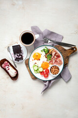 Traditional Englis breakfast plate with bacon strips, sunny side up eggs, vegetables and cake on light background