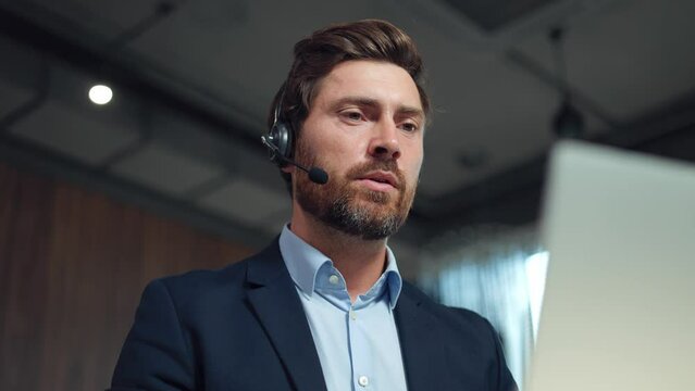 Low angle view of serious bearded man in stylish suit wearing wireless headset sitting at desk with modern laptop and talking during video chat. Concept of business, technology and communication.