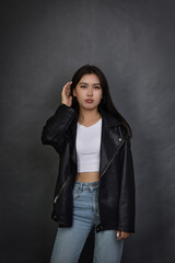 Asian girl in modern clothes stands against gray wall, straightening hair with hand. Brunette with long hair in leather jacket and jeans raised hand up, looking wistfully straight at the camera