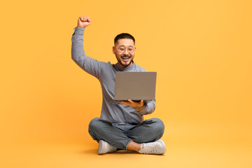 Joyful Asian Man Celebrating Success With Laptop While Sitting On Yellow Background