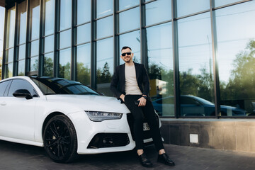 Portrait of rich businessman wearing suit, standing near his luxury black car and using smartphone...