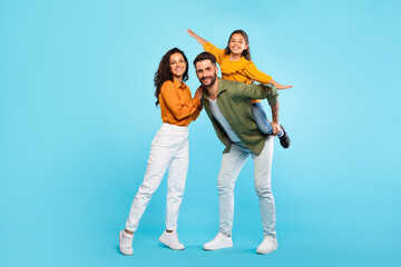 Cheerful european family of three having fun, dad piggybacking daughter over blue background, full...