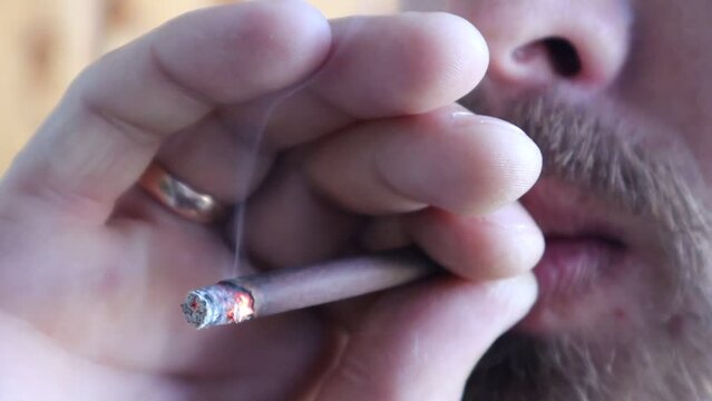 A close-up on a small cigar. A man smokes a cigar, it smolders, then he releases the smoke. Smoking is a bad habit. High quality video