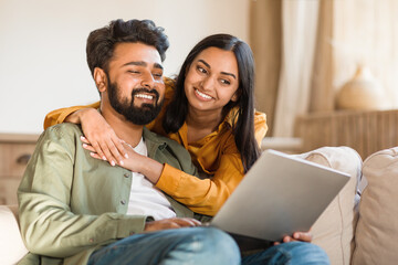 Happy young indian couple using laptop at home together, shopping online or booking vacation,...