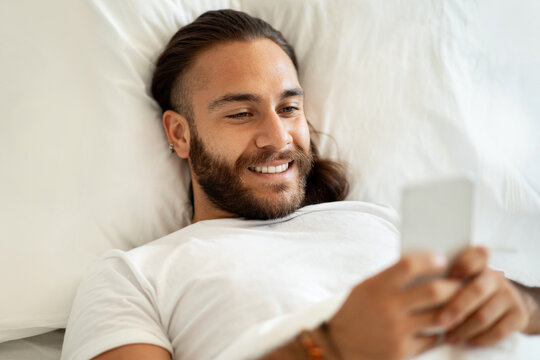 Happy young long-haired man lying in bed, using smartphone