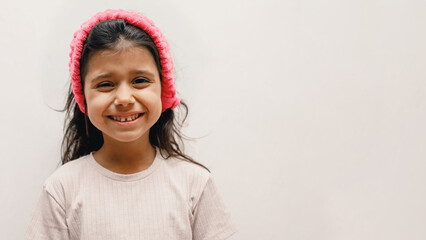 Cheerful Spanish girl child in a beige T-shirt and a pink headband on a white background, she shows...