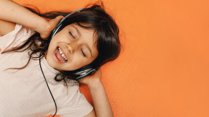 Spanish girl with dark hair listens to music on headphones inside the house, orange background, close-up
