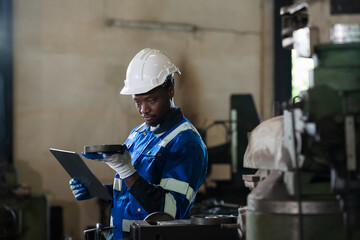 Male engineer worker using digital tablet during working with lathe machine in industry factory, wearing safety uniform, helmet. Male technician worker maintenance parts of machine in workshop plant