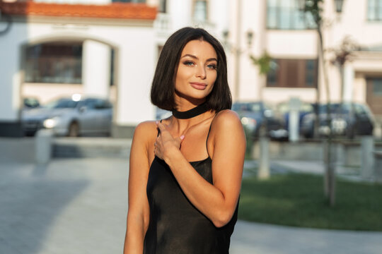 Beautiful fashion young fresh summer girl with short hairstyle in an elegant black spaghetti strap dress walking in the city at sunset. Pretty chic adorable woman