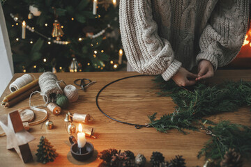Making Christmas rustic wreath. Hands holding cedar branches, making wreath on wooden table with pine cones, candle, twine, bells in atmospheric festive room. Winter holiday preparations