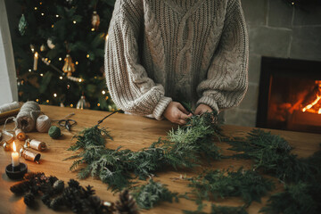 Making Christmas rustic wreath. Hands holding cedar branches, making wreath on wooden table with pine cones, candle, twine, bells in atmospheric festive room. Winter holiday preparations