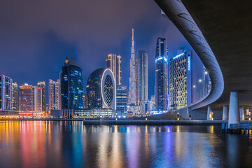 Dubai at night. City skyline in United Arab Emirates. Skyscrapers illuminated at the blue hour with a curved course of a bridge. Reflections on the water surface in the port - obrazy, fototapety, plakaty