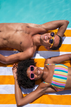 Smiling friends lying on stripped towel with eyes closed near pool