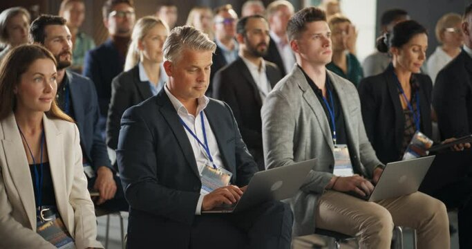 Caucasian Male Pharmaceutical CEO Sitting In Diverse Crowd At Medical Summit And Using Laptop Computer. Successful Man Listening To Presentations About New Developments In Biopharma and Medicine.