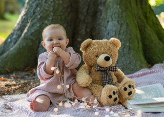 Portrait of an adorable little girl in a beige dress sitting near a large tree and looking at the...