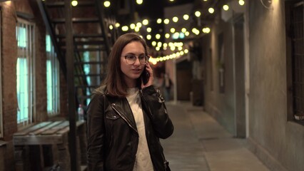 A young woman talks on the phone on a narrow street in the evening. A girl with glasses and a leather jacket on a cozy street.