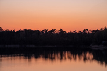 Nature's breathtaking artistry unfolds as a serene forest lake mirrors the dramatic transformation of the sky from vivid blue to fiery orange during dusk. The tranquil waters capture the essence of