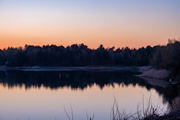 Nature's breathtaking artistry unfolds as a serene forest lake mirrors the dramatic transformation of the sky from vivid blue to fiery orange during dusk. The tranquil waters capture the essence of