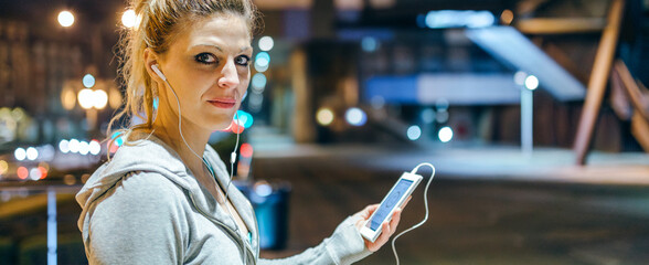 Portrait of young blonde woman looking at camera while listening music on mobile phone application. Banner of female runner with earphones holding cell with heart rate display after training at night
