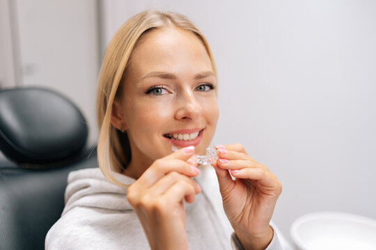 Closeup portrait of smiling blonde female patient holding invisible braces aligner sitting on chair in dentistry clinic. Close up of mobile orthodontic appliance for dental correction.