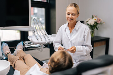 Front view of smiling female dentist doctor having consultation with unrecognizable little child girl lying on dental chair in dentistry clinic. Concept of children teeth treatment, pediatric checkup.