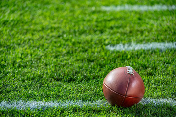 A low angle close-up view of a leather American Football sitting in the grass. It is on a white yard line with hash marks in the background.