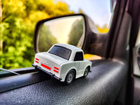 Small white toy car on background of side mirror of a real big caron and nature landscape. Concept of travel and adventure by car. Freedom of movement on your own transport. Blur and partial focus