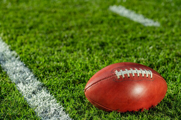 A low angle close-up view of a leather American Football sitting in the grass. It is next to a white yard line with hash marks in the background. - obrazy, fototapety, plakaty
