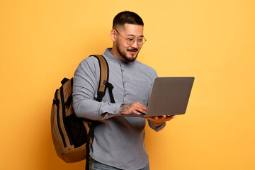 Young Asian Student Guy With Laptop And Backpack Standing On Yellow Background