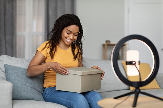 Happy Black Female Blogger Unpacking Cardboard Box, Shooting Video On Phone Set On Ring Lamp, Sitting In Living Room