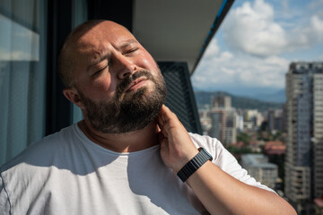 Bearded man in hot summer sun on balcony. Bald, unsmiling guy closed eyes in displeasure discontent from discomfort, holding on to sore neck, kneading ligaments, suffering from protrusion, pain, ache - obrazy, fototapety, plakaty