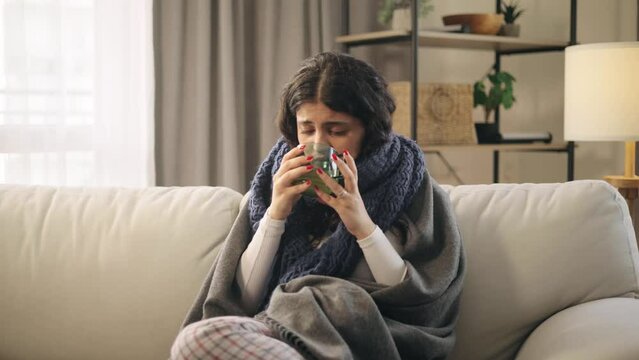 Sick curly woman sitting on sofa under the warm blanket coughing having severe headache temperature and drinking hot tea at home Unhealthy female getting flu fever virus symptom indoors