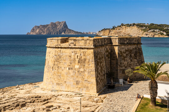 A panoramic view of the historic stone Moraira Castle on the rocky coast, with the prominent Pe&ntilde;on de Ifach rock formation rising in the distance across the blue sea, Alicante, Spain.