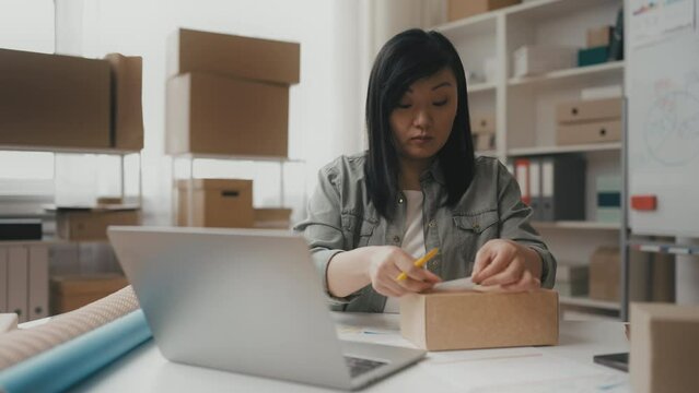 Middle-aged Asian woman entrepreneur putting sticker on box, packing orders