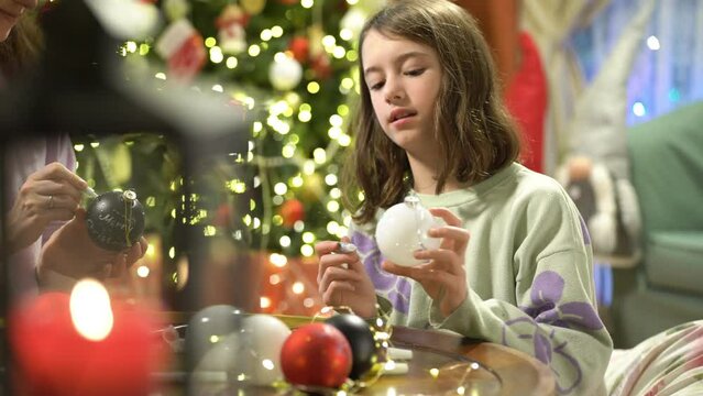 A young attractive mother and her cute daughter are painting Christmas decorations on Christmas Eve, preparing for the celebration and decorating the Christmas tree