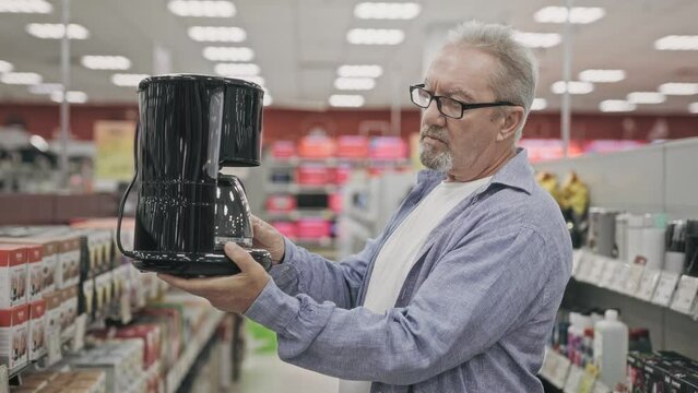 An elderly man in an electronics hypermarket chooses a coffee maker. A pensioner buys a coffee machine in a shop