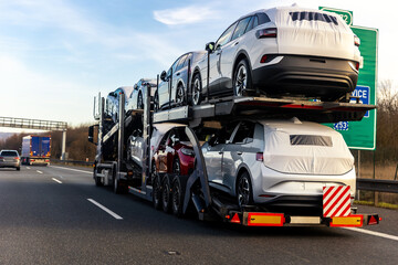 Tow truck car carrier semi trailer on highway carrying batch of new wrapped electric SUVs on motorway road at sunset evening time. Business distribution logistics service. Lorry driving highway - obrazy, fototapety, plakaty