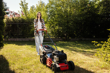 Female gardener working in autumn, cutting grass in backyard. Concept of gardening, work, nature. Housework, gardening and country life. Home garden grass cutting woman mowing with lawn mower.