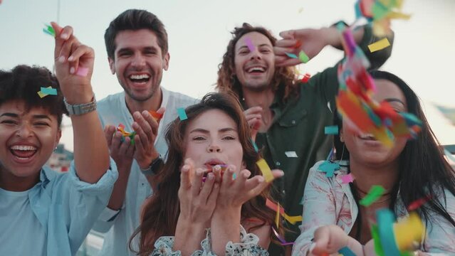 Cheerful multicultural friends blowing up holiday clapper while celebrating birthday on restaurant balcony