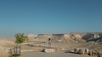 European woman stands against the background of the Negev Desert in Israel is visible in the background over mountains, sandy hills and rock formations.