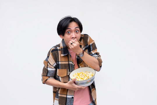 A young wide-eyed asian man voraciously stuffing his mouth with popcorn while watching a movie. Isolated on a white background.