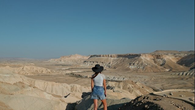 European woman stands against the background of the Negev Desert in Israel is visible in the background over mountains, sandy hills and rock formations.