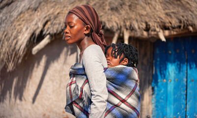 african mother in the village carry her child in the back in a traditional way in a blanket,...