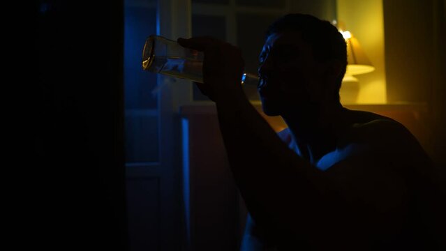 Silhouette of a Depressed Man Drinks Alcohol from a Glass Bottle at Night in the Dark. Problems with Alcoholism and Drug Addiction. Slow Motion.