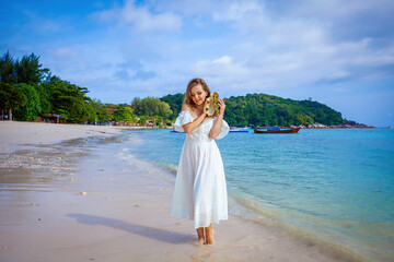 Fototapeta na wymiar Woman in white dress holds seashell, enjoys the ocean view. Concept of beach holiday and leisure.