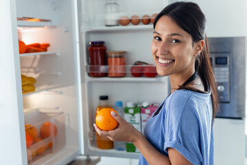 Beautiful young woman taking an orange from the fridge while looking at camera in the kitchen at...