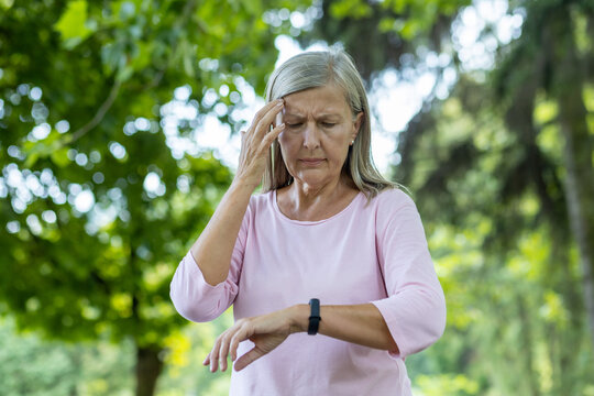 An Older Gray-haired Woman Feels Bad After Jogging And Active Sports. Standing In The Park, Holding His Head And Looking At The Smart Watch, Checking His Pulse And Breathing.