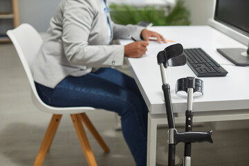 Disabled woman in an office workplace. Orthopedic elbow crutches by an office desk, with a disabled...