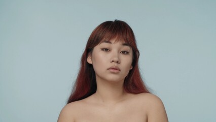 Close-up isolated photo of a young seminude woman with red dyed hair and nude makeup looking straight at the camera with a relaxed look on her face.